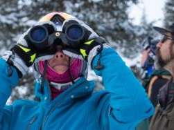 A woman in a blue coat stands in a snowy landscape and holds binoculars to her eyes.