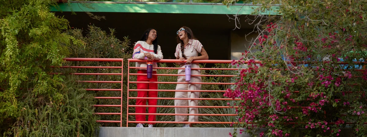 Two people stand on a vine-covered balcony while holding beautiful Stanley water bottles